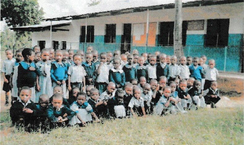 A large group of young pupils in blue and white uniforms gathered on the grass in front of a classroom block