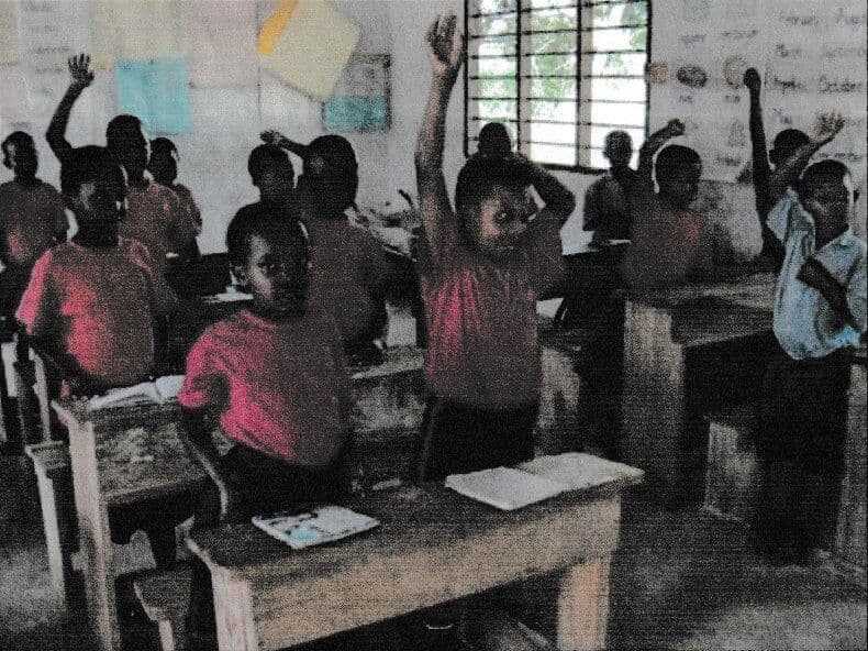 Pupils in red T-shirts standing at wooden desks in a classroom, several with hands raised