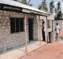 People standing beside a new stone classroom block with a tin roof and veranda posts, palm trees behind