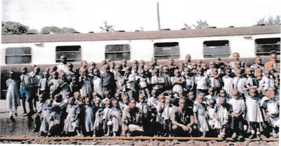 A large group of pupils and teachers posing beside a passenger train on the tracks