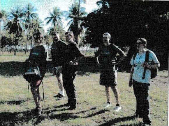 Five volunteers with backpacks standing on grass among palm trees