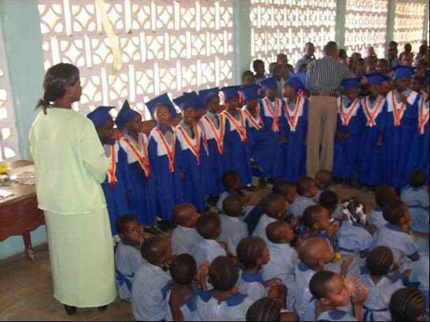 Kindergarten graduates in blue gowns, mortarboards and sashes standing in a row, with pupils seated on the floor in front