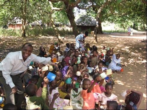 A smiling man in a white shirt crouching among a large group of children holding plastic cups under shady trees