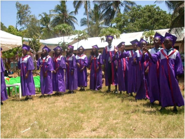 Graduating pupils in purple gowns and mortarboards standing in a line on the grass in front of a school building