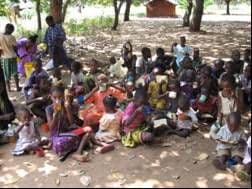 Children and adults sitting on the ground in the shade of trees, receiving food