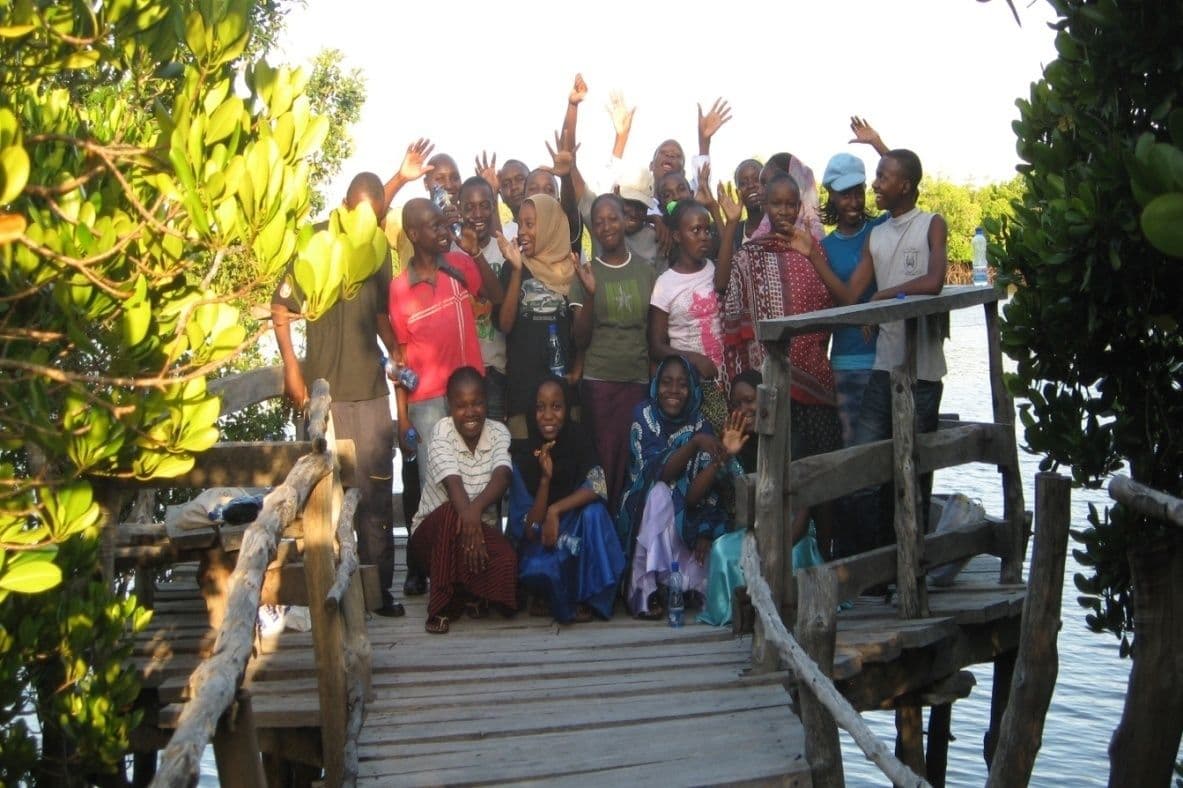Standard 8 graduates waving on a wooden boardwalk among mangrove trees by the water