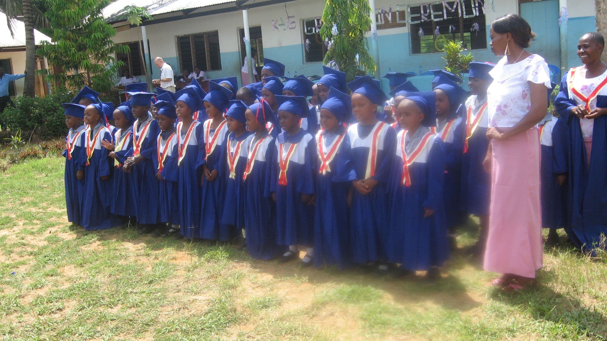 Rows of nursery children in blue graduation gowns and caps standing outside a classroom with two teachers