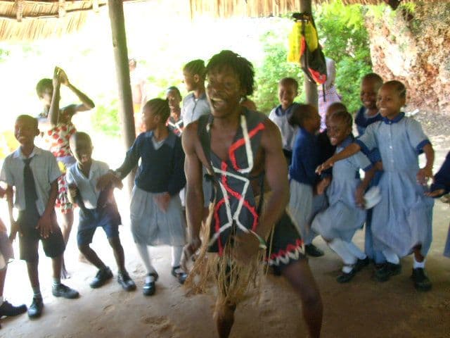A performer in traditional costume dancing with laughing pupils under a thatched shelter