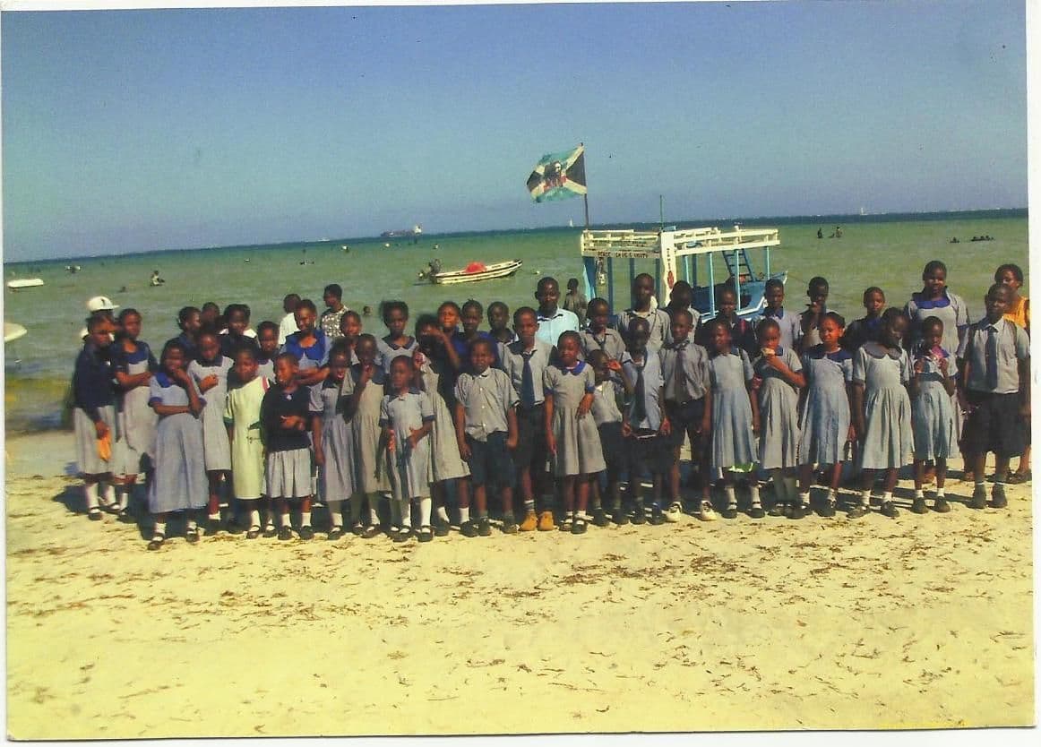 Pupils in grey and blue school uniform standing on a sandy beach with boats and the sea behind