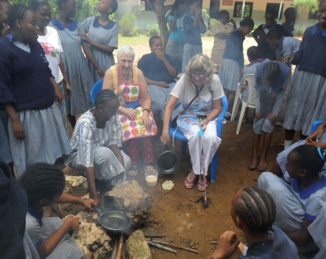 Two visiting women seated among pupils gathered round a charcoal stove cooking outdoors