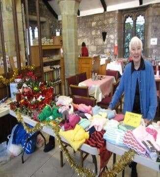 A woman beside a stall of knitted clothes and Christmas gifts in a church hall