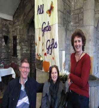 Three fayre helpers smiling in front of a banner reading All God's Gifts