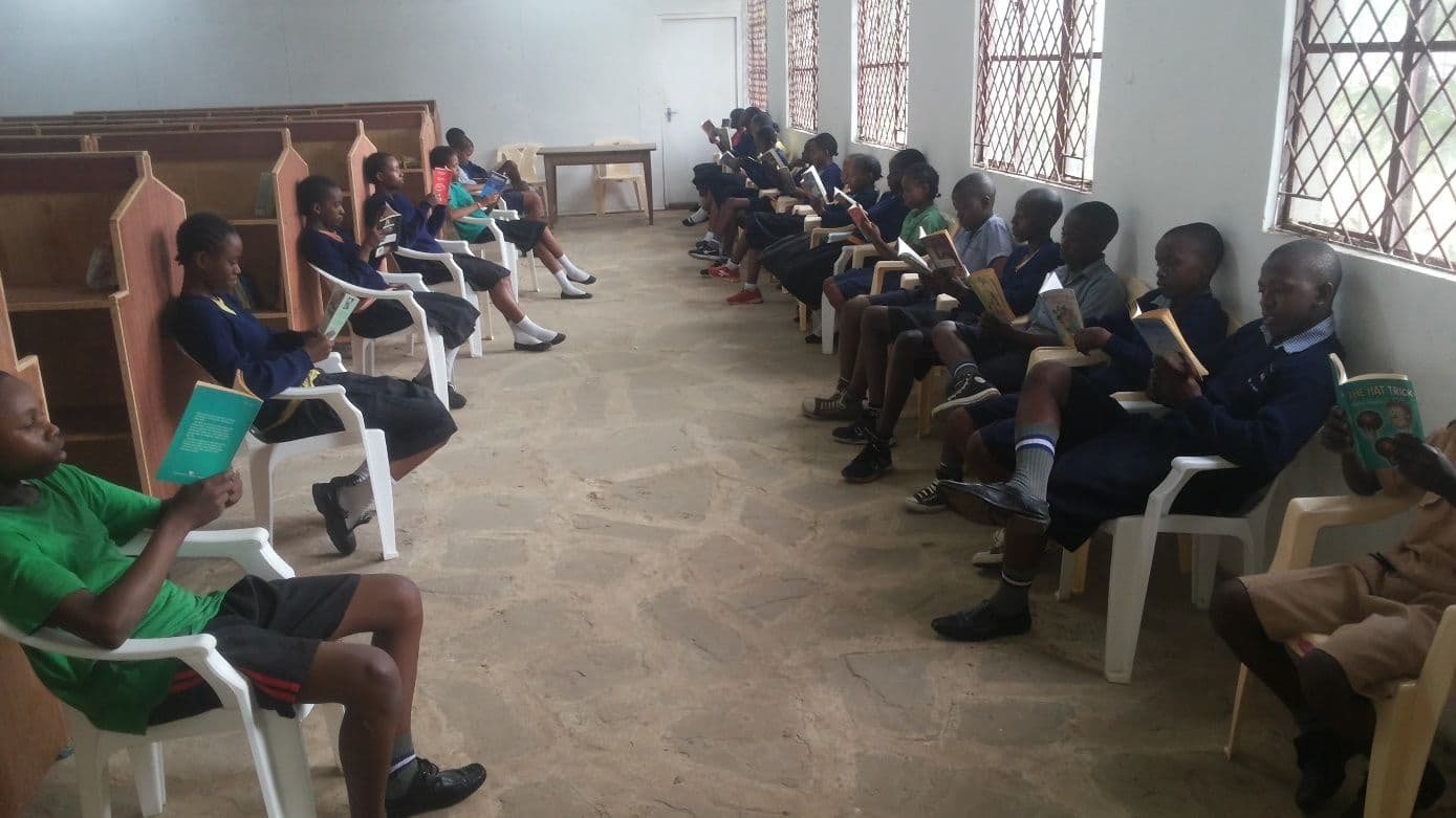 Pupils in school uniform sitting in two rows of chairs reading books in the library
