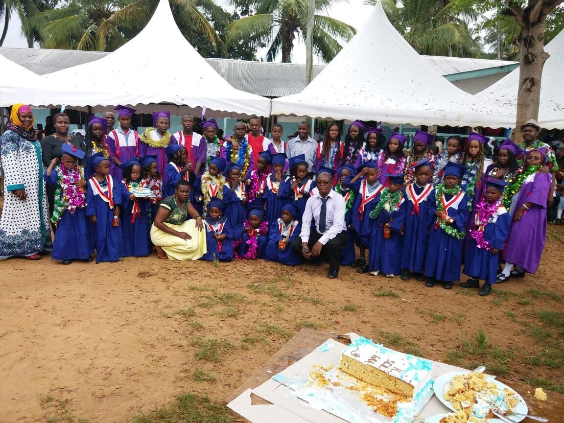 Graduating pupils in purple and blue gowns with garlands, posed with teachers under white tents