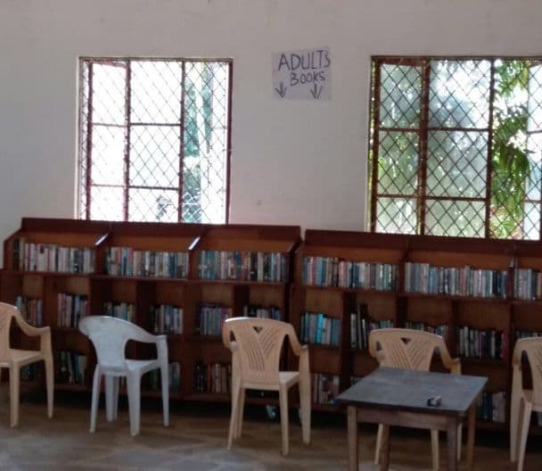 Library shelves of books under an "Adults books" sign, with plastic chairs and a table