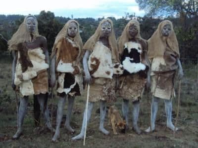 Five young men covered in white clay and wearing animal-skin capes standing on grassland