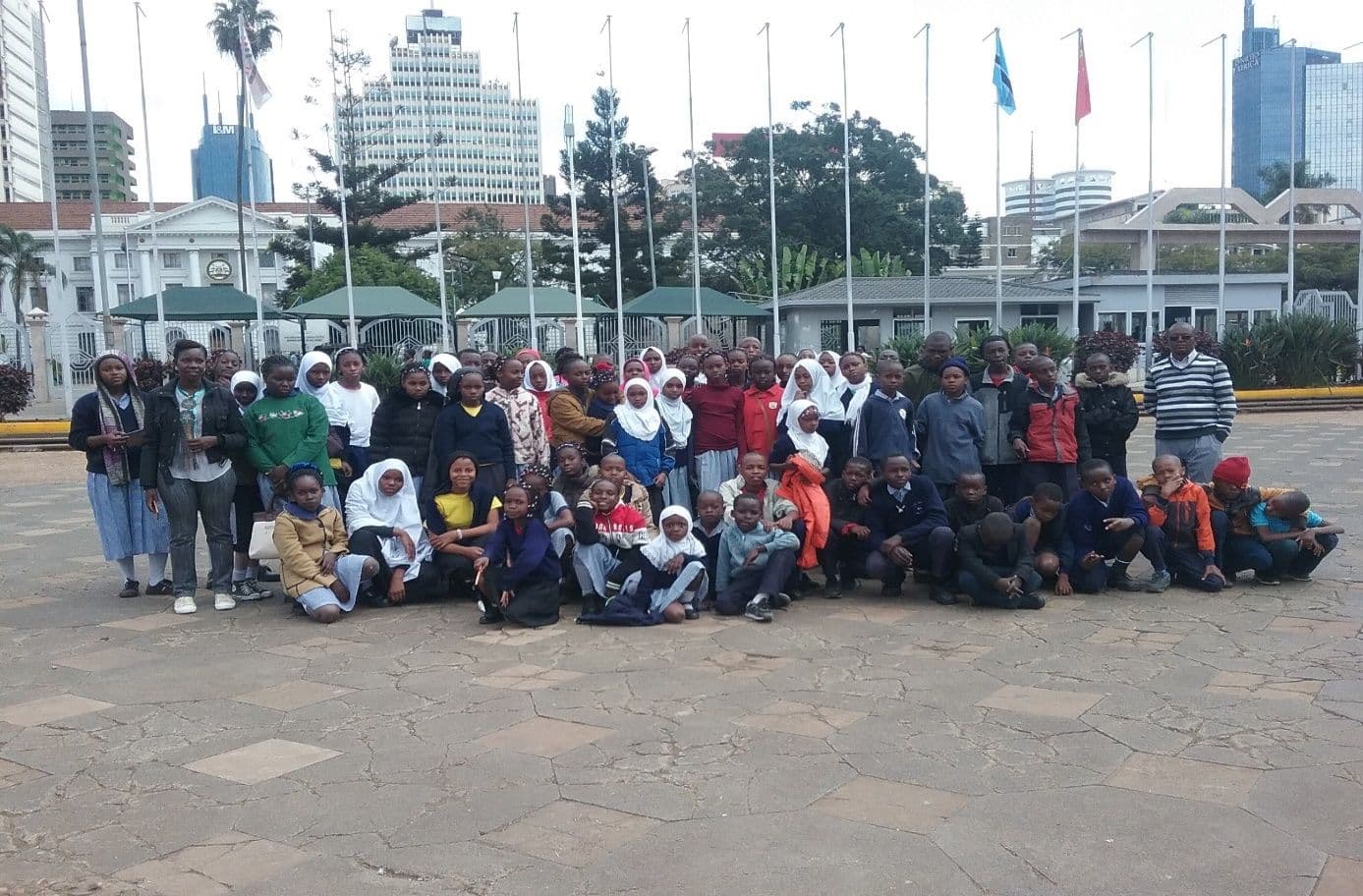 Large group of pupils and teachers posing on a paved plaza with flagpoles and city tower blocks behind