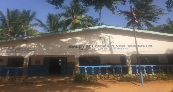 School building painted white and blue with the Emley Education Centre Msambweni sign, under palm trees