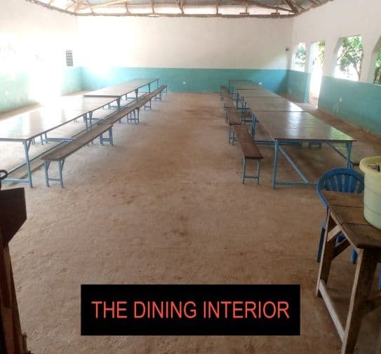 Empty dining hall with long tables and benches on a sandy floor under an iron roof