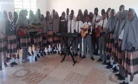 Students in uniform standing in rows with guitars and a keyboard, a choir in a classroom