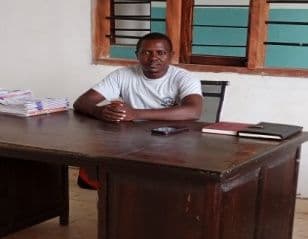 Man in a white T-shirt seated at a wooden office desk by a window