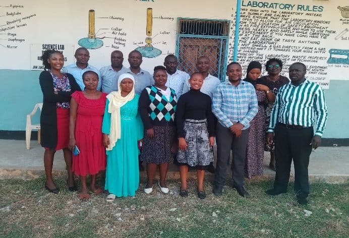 Teaching staff, men and women, standing in two rows outside the science lab wall painted with lab rules