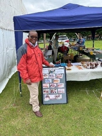Man in a red jacket beside a photo display board at an outdoor show stall under a blue gazebo