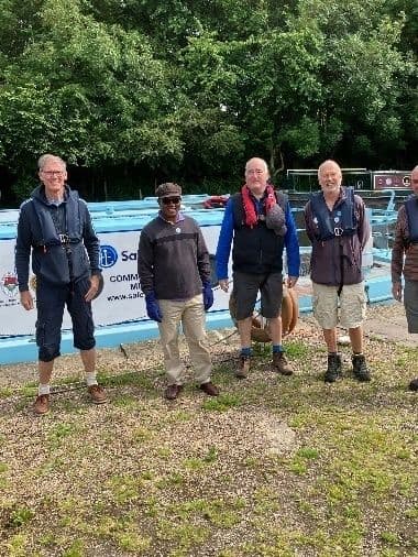 Group of men, several in life jackets, standing on gravel in front of a canal boat