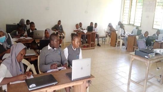 Students in grey school uniforms working on laptops at wooden desks in the computer lab