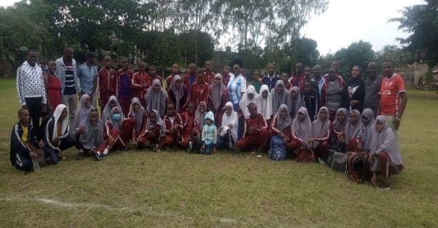 Large group of students in maroon tracksuits and headscarves with coaches, posing on a grass field