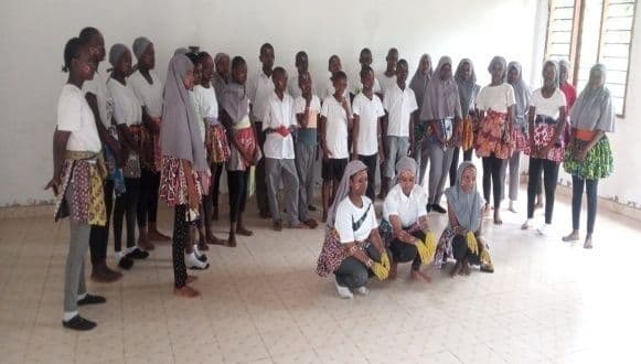 Students in white tops and African-print wraps rehearsing a dance in a hall, three kneeling in front