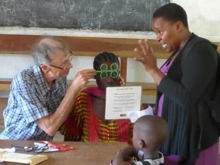 A man holds trial lens frames to a woman's eyes as she reads a test card in a classroom