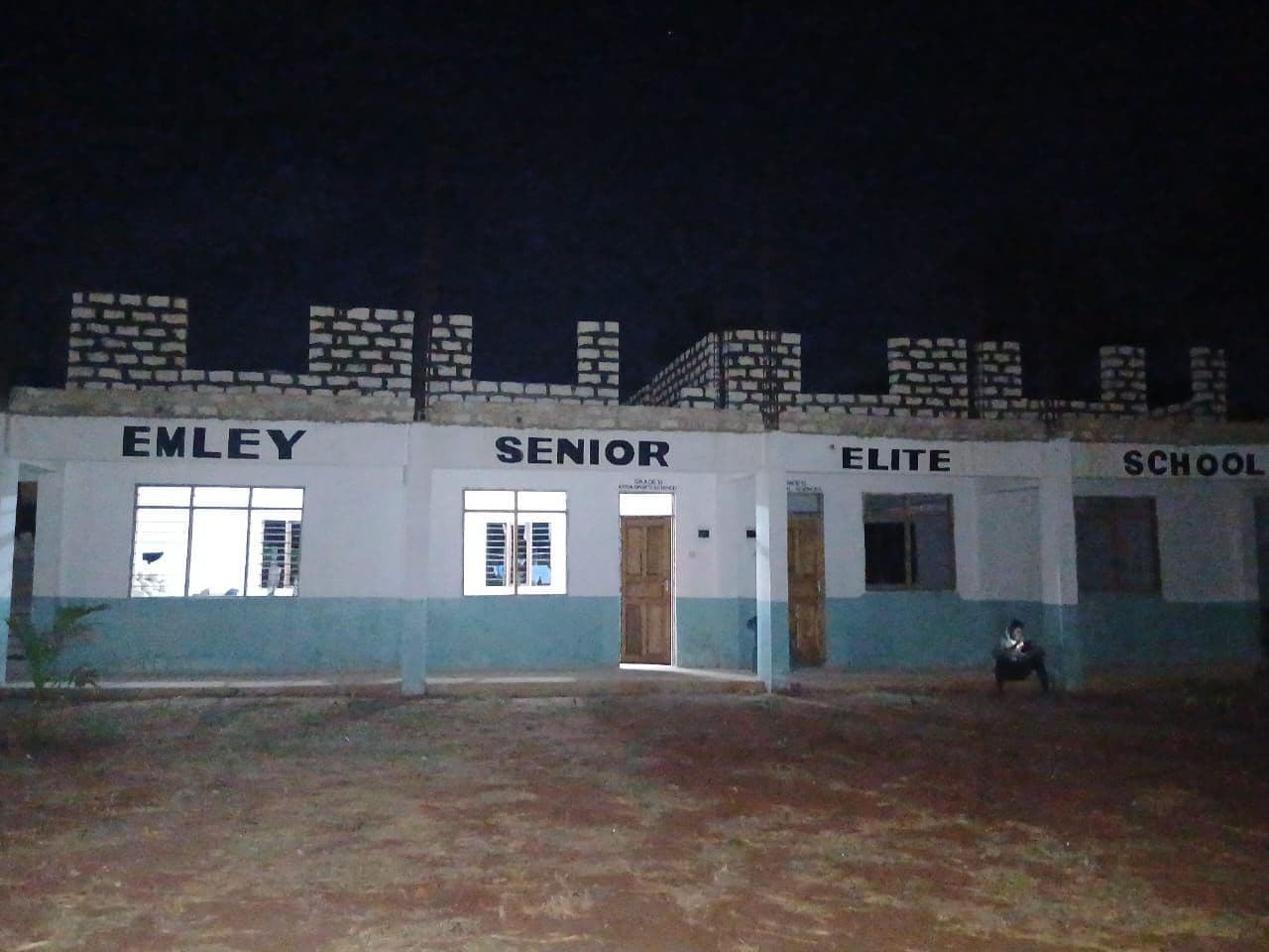 Night view of a lit single-storey building painted EMLEY SENIOR ELITE SCHOOL, with block walls rising above