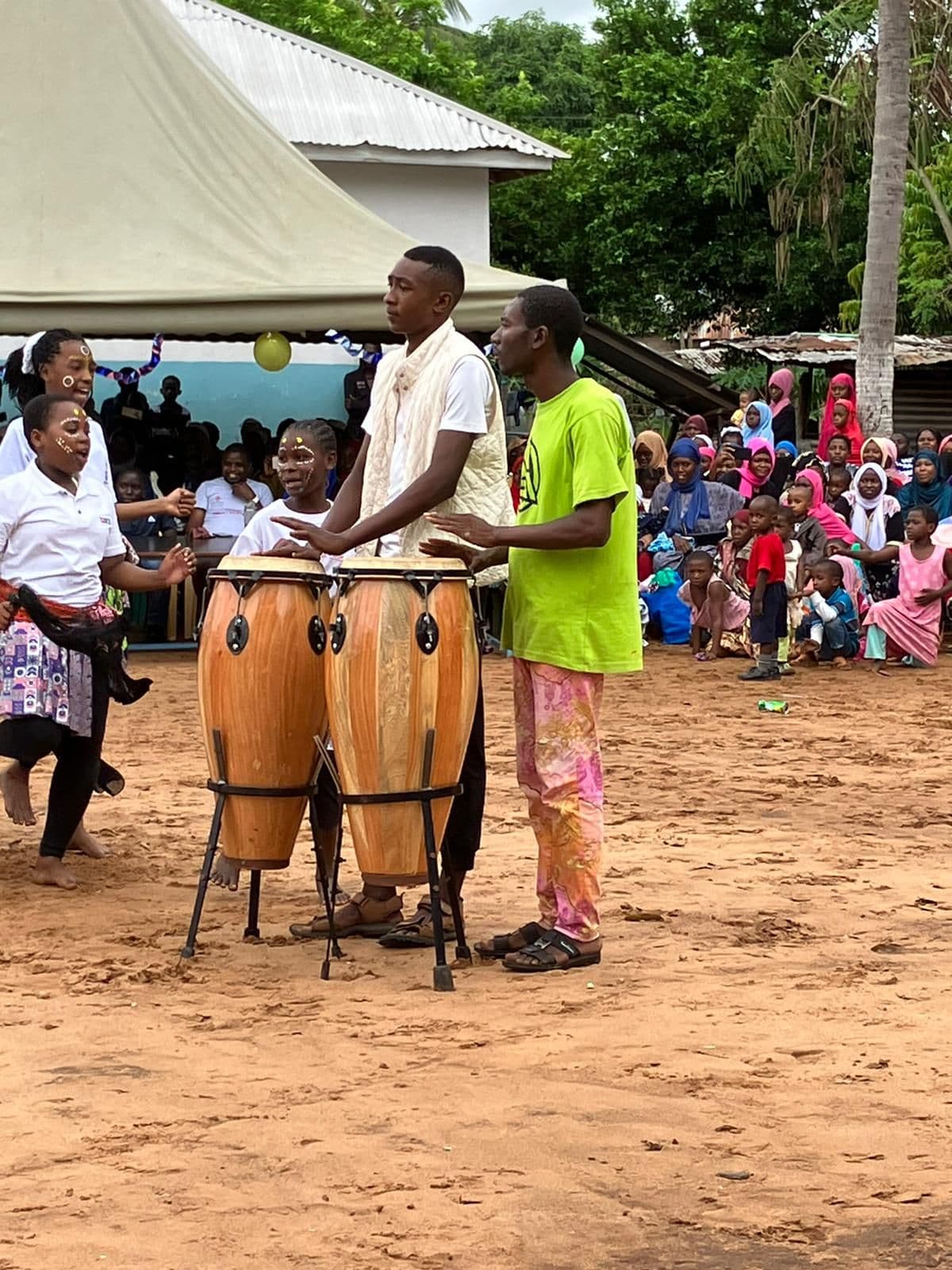 Two men playing tall drums in the yard as a crowd watches from under shelter
