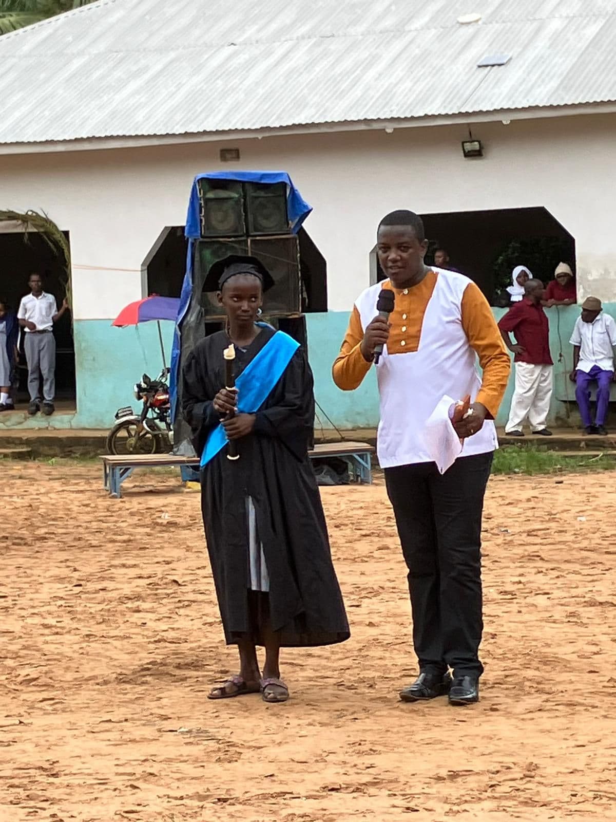 A pupil in a black gown and sash stands beside a man with a microphone