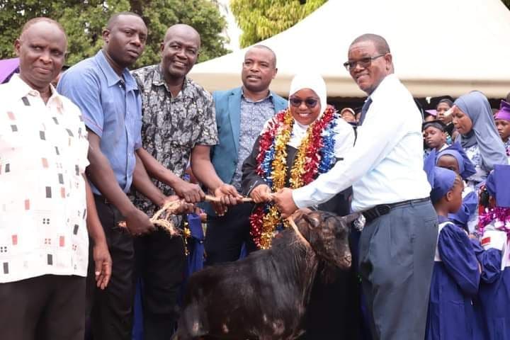Men and a woman in tinsel garlands holding a goat on a rope among pupils in gowns