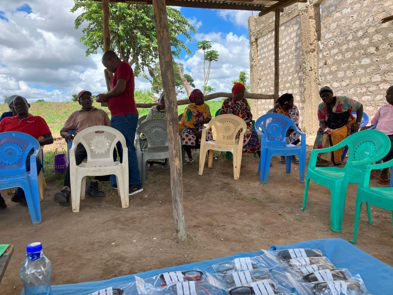 Villagers seated on plastic chairs under a shelter with spectacles laid out on a blue cloth