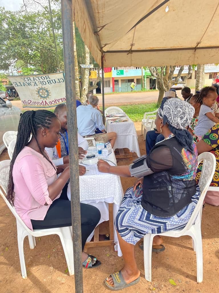 People seated under a tent at tables during an outdoor medical camp