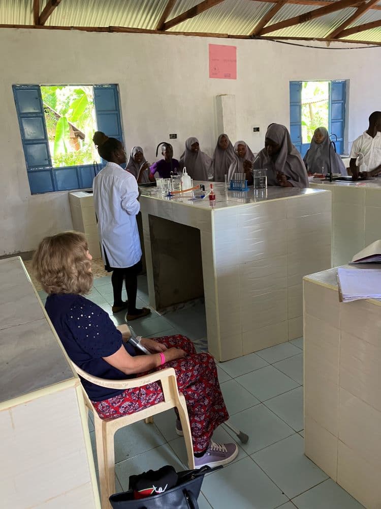 A visitor sits watching as a teacher in a lab coat leads pupils at lab benches