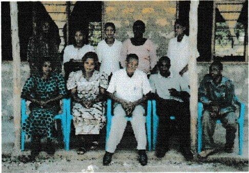 Teachers posing on a veranda, five seated on blue plastic chairs and five standing behind them