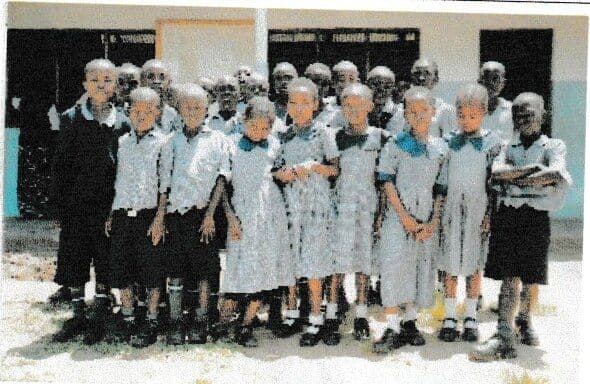 About twenty pupils in grey-and-white uniforms standing in rows in front of a classroom