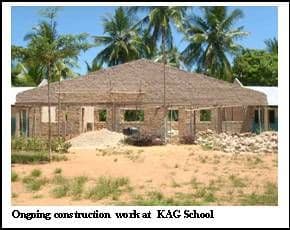 Unfinished brick school building with an iron roof frame, surrounded by palm trees and building stones