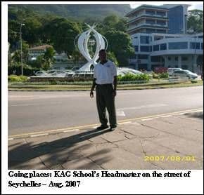 A man in a white shirt standing on a street in Seychelles, with a sculpture and buildings behind him