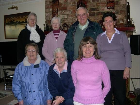 Seven fundraisers, six women and one man, posing in two rows in a living room in front of a brick fireplace