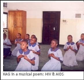 Girls in school uniform kneeling on a classroom floor with hands clasped, performing a choral verse