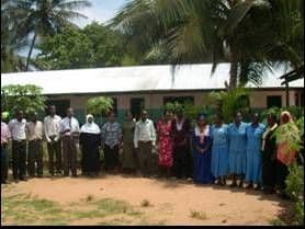 About twenty teachers standing in a line in front of a school building with palm trees behind
