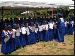 Graduating pupils in blue gowns and caps holding certificates, lined up under a white tent