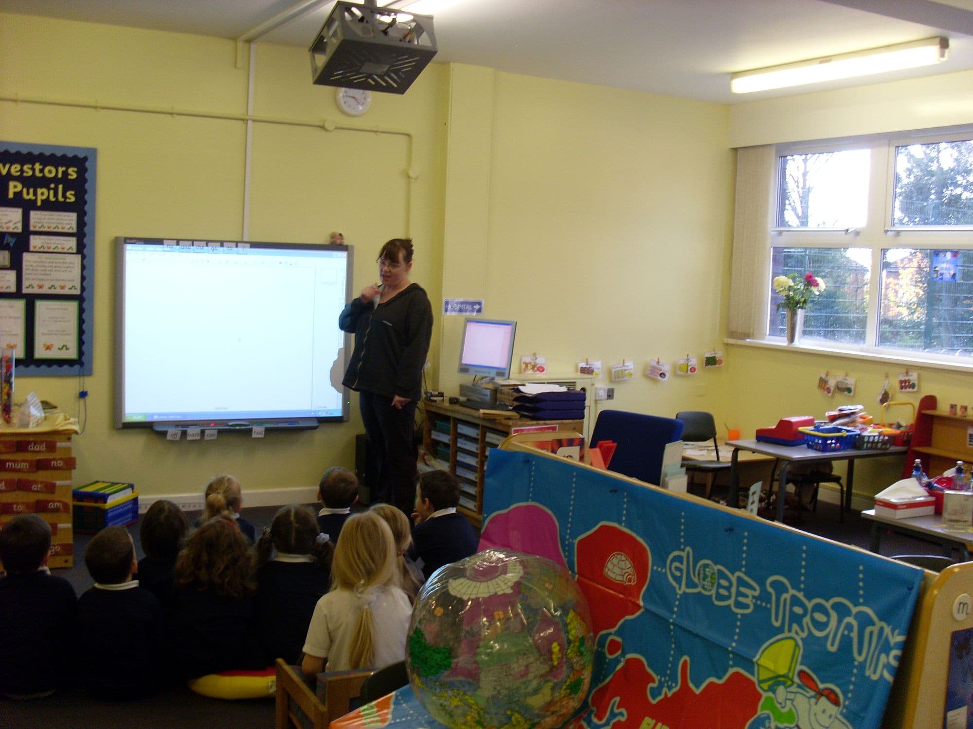 A teacher standing at an interactive whiteboard while young pupils sit on the carpet in a UK classroom