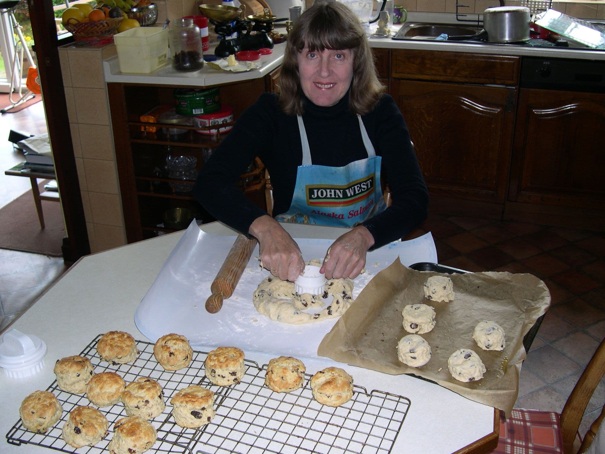 A woman in an apron cutting scones from dough at a kitchen table, with baked scones cooling on a rack