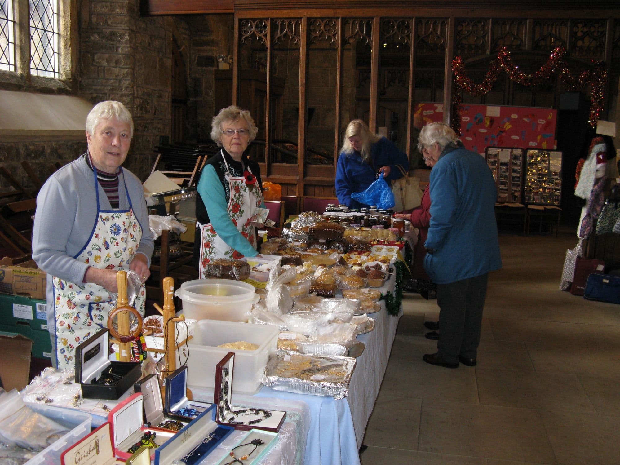Two women in aprons behind a long table of cakes, jams and jewellery at a church fayre, with shoppers browsing